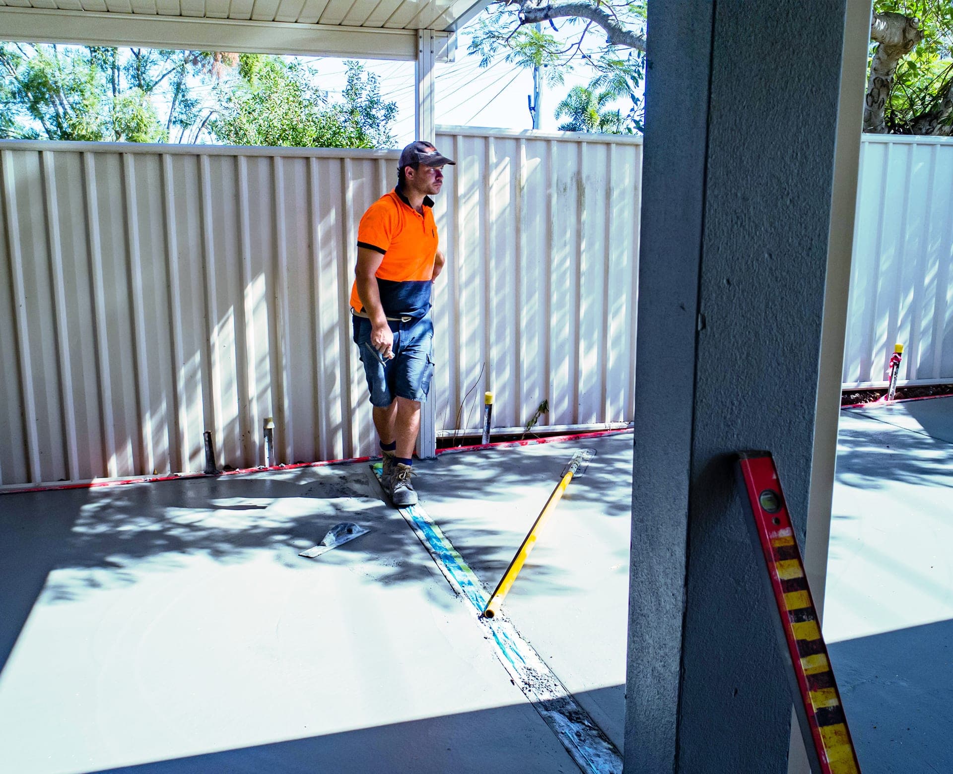 A concreter in a high-vis polo and cap on a freshly laid concrete carport slab, with a float and a spirit level nearby.