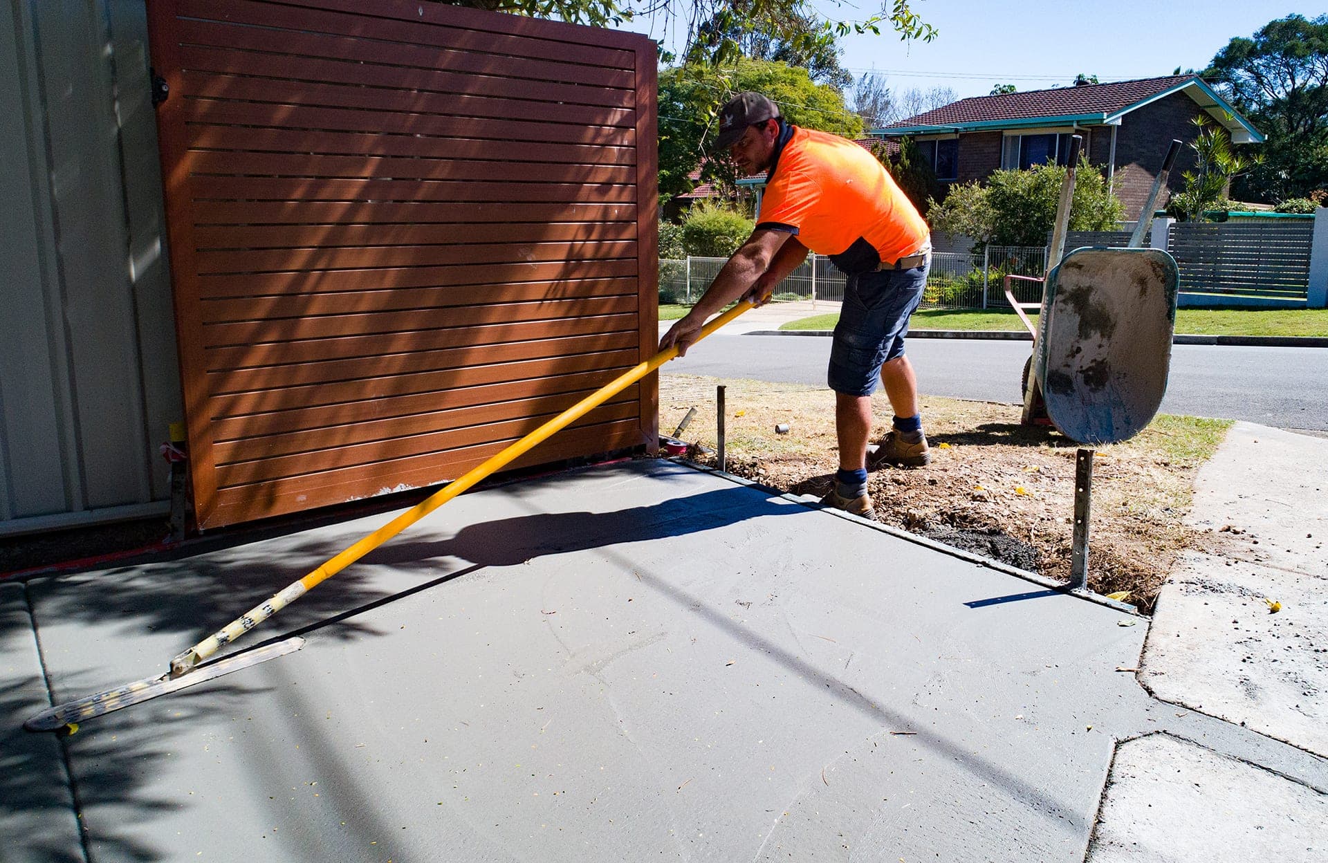 A concreter pulling a long-handled float across a freshly poured concrete slab beside a timber gate.