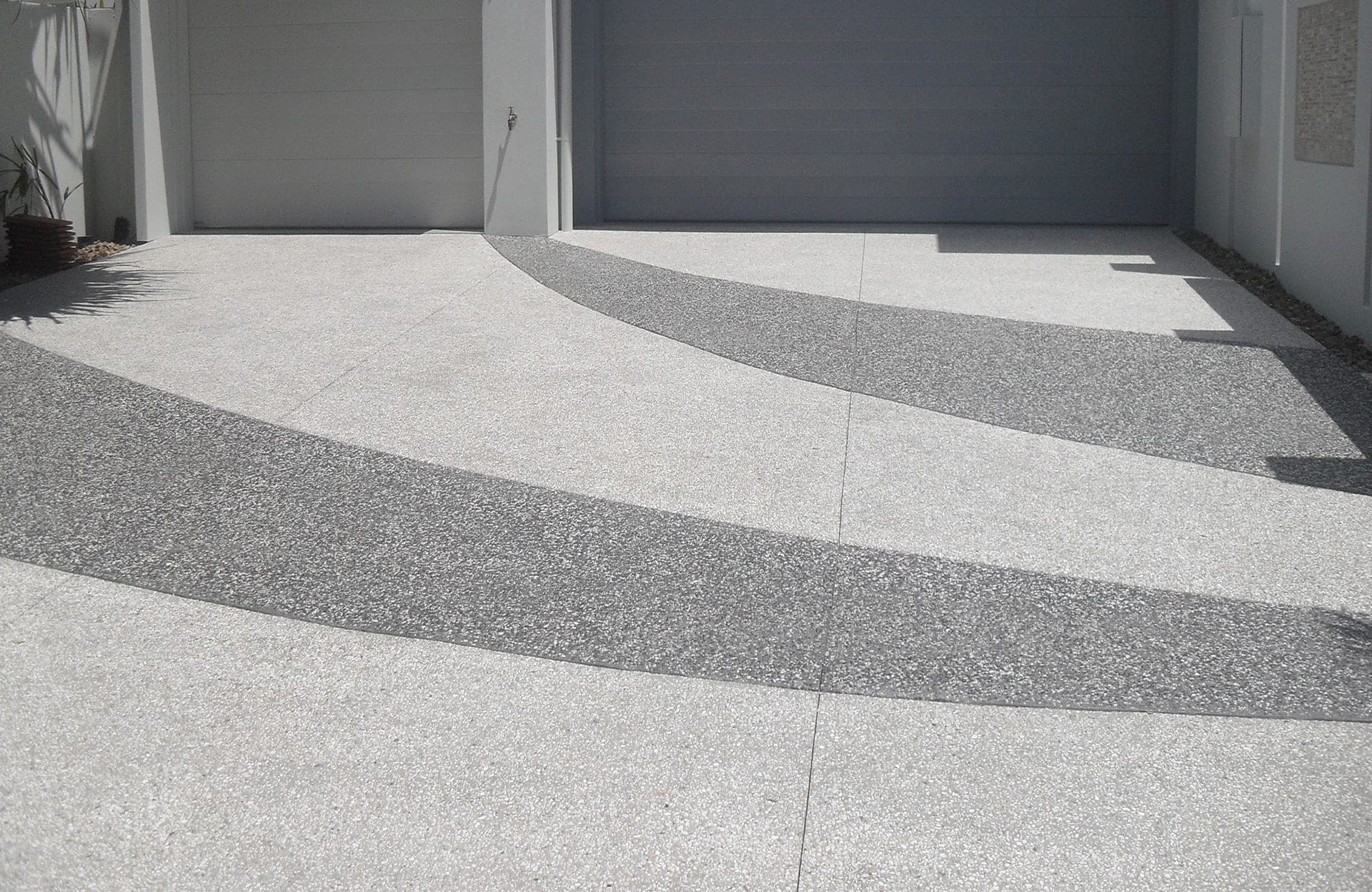 New exposed aggregate concrete driveway in front of two garage doors, with two broad curved bands of darker aggregate sweeping across the lighter slab.