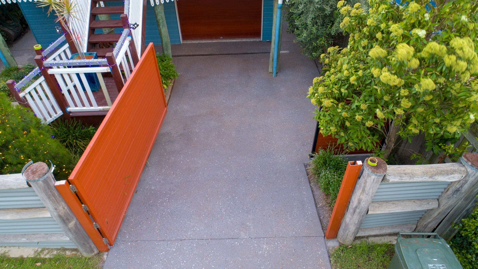 Large-drop Grey Covercrete driveway with light flecks, running from an open orange gate to the garage under a raised weatherboard house.
